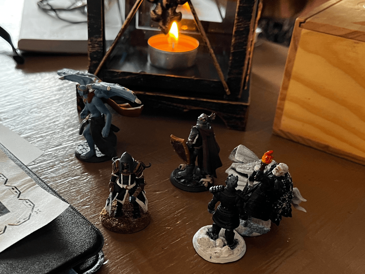 Miniature figures on a table: dragon, cloaked figures, and a lantern with a lit candle in the background.