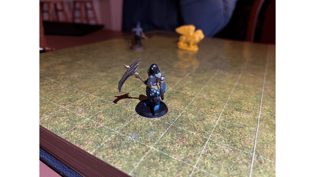 Miniature of a figure with a scythe on a grid board, with two other miniatures visible.
