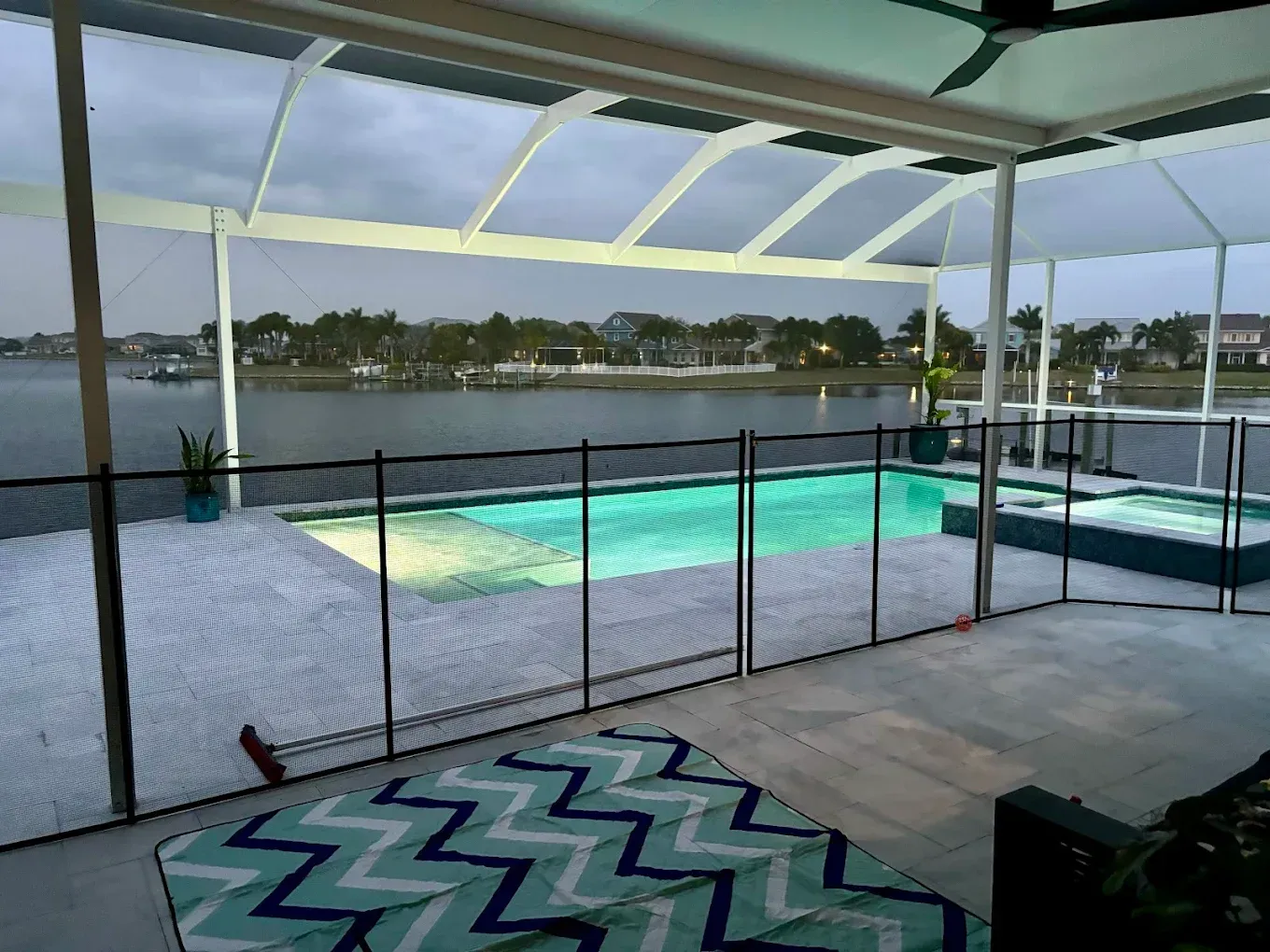 Pool area with black fence, overlooking water. White screened-in enclosure, blue rug, overcast sky.