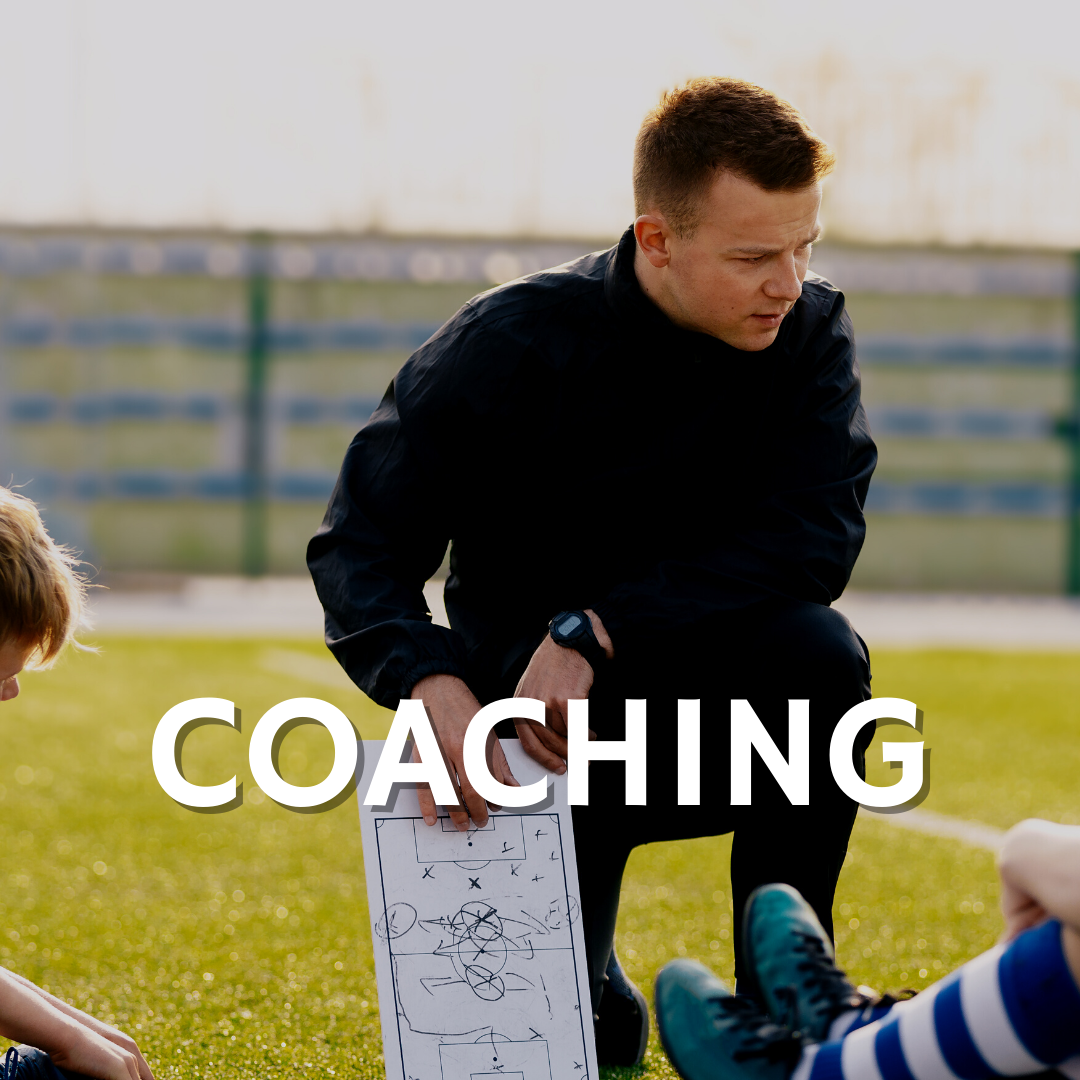 A man is kneeling down on a soccer field holding a white board with the word coaching on it