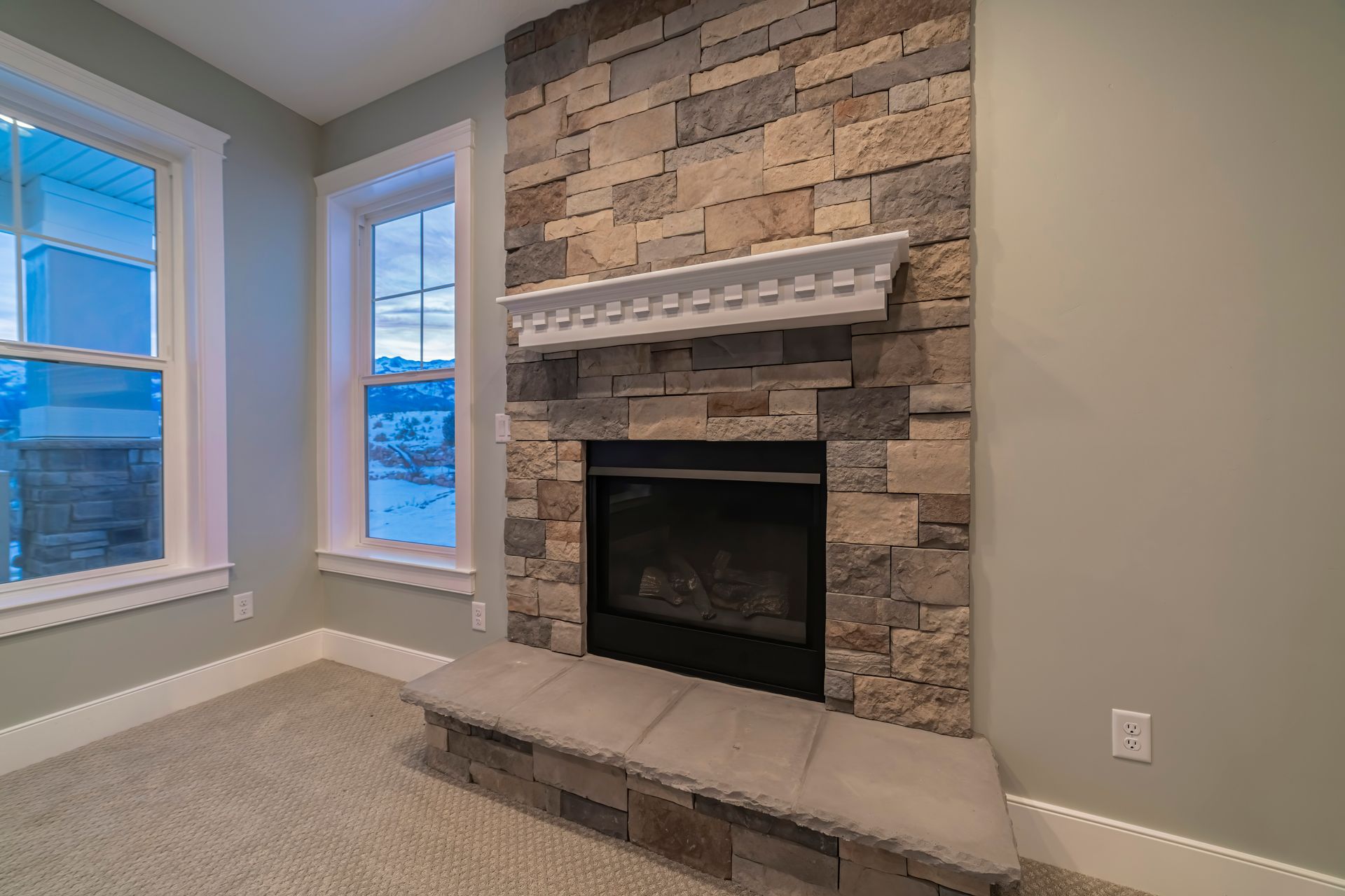 Fireplace with stone surround, mantle, and cushioned bench, flanked by windows in a light-colored room. Fireplace with stone surround, mantle, and cushioned bench, flanked by windows in a light-colored room.