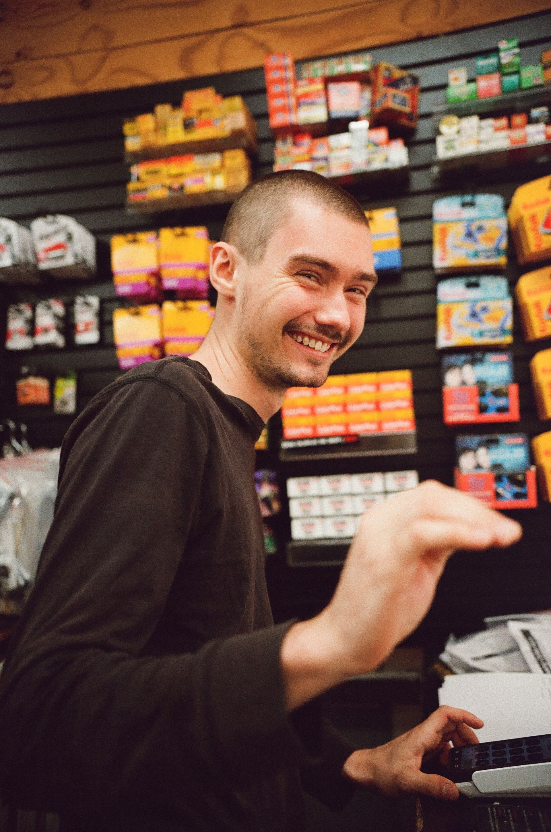 Young woman with dark hair and a plaid shirt smiles broadly inside a store, near a refrigerator.