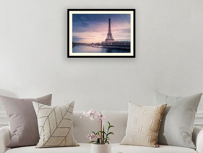 Eiffel Tower framed above a couch with pillows and a small potted plant. Soft pink and blue hues in the sky.