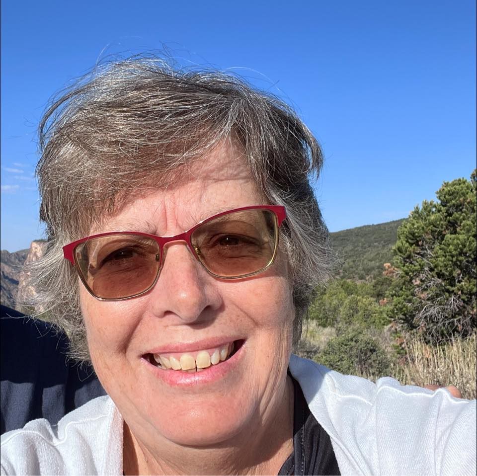 Woman with glasses smiles outdoors, with a mountain backdrop.