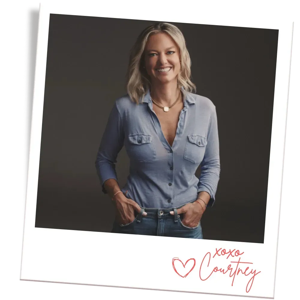 Woman in blue shirt smiles, poses hands in pockets. Polarid-style photo with a signature.
