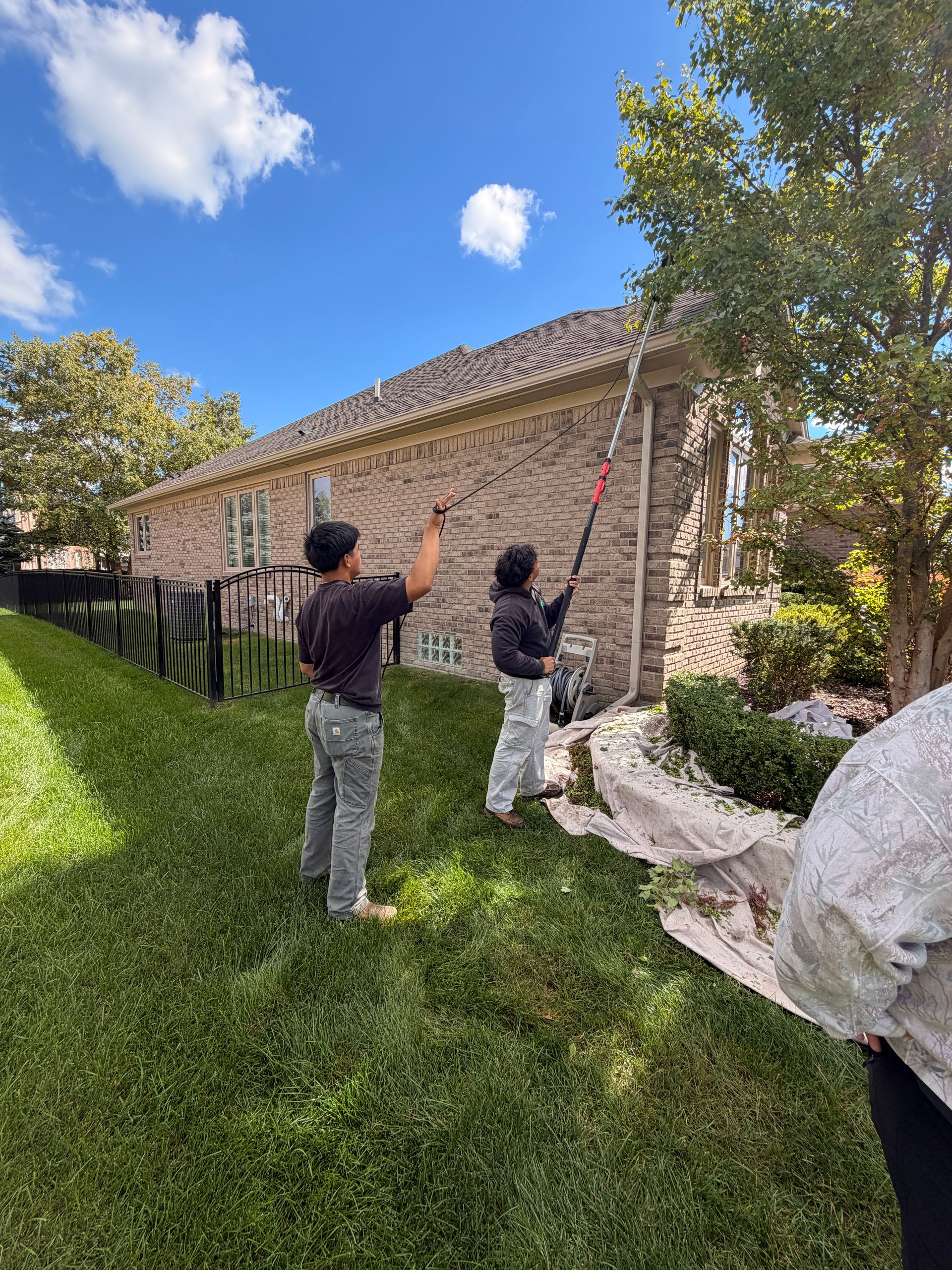 Workers painting a brick house exterior beside a grassy yard on a sunny day