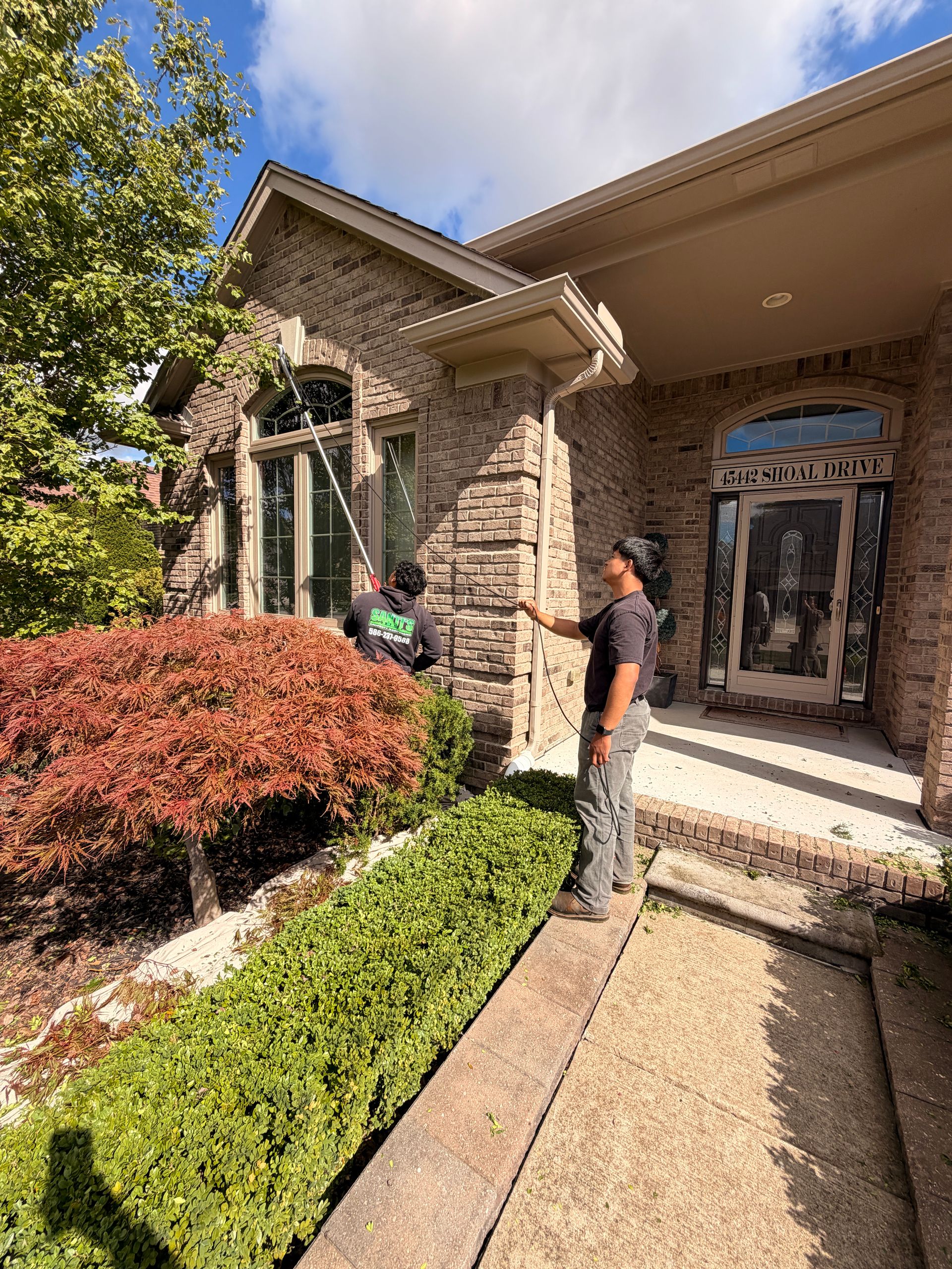 Two people standing outside a stone house with a front walkway, shrubs, and arched entryway.