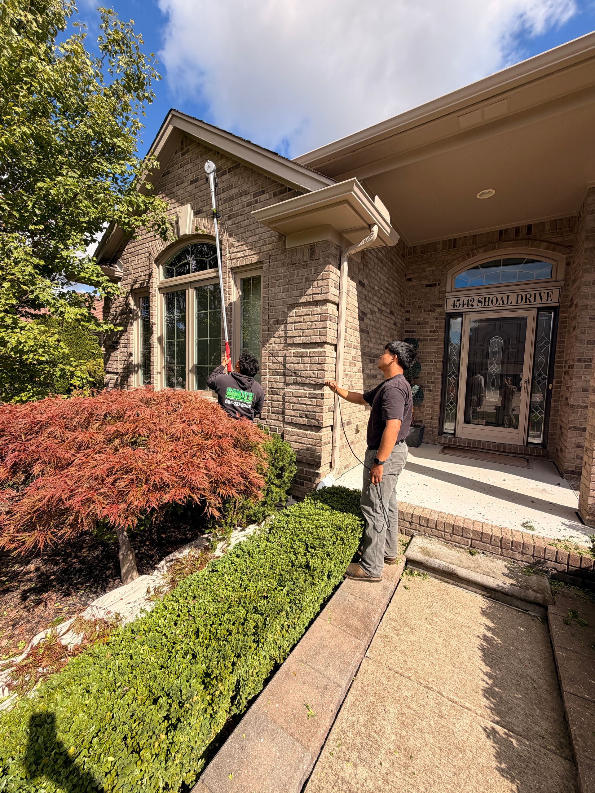 Person standing outside a stone house with arched windows, near trimmed hedges and a front walkway.