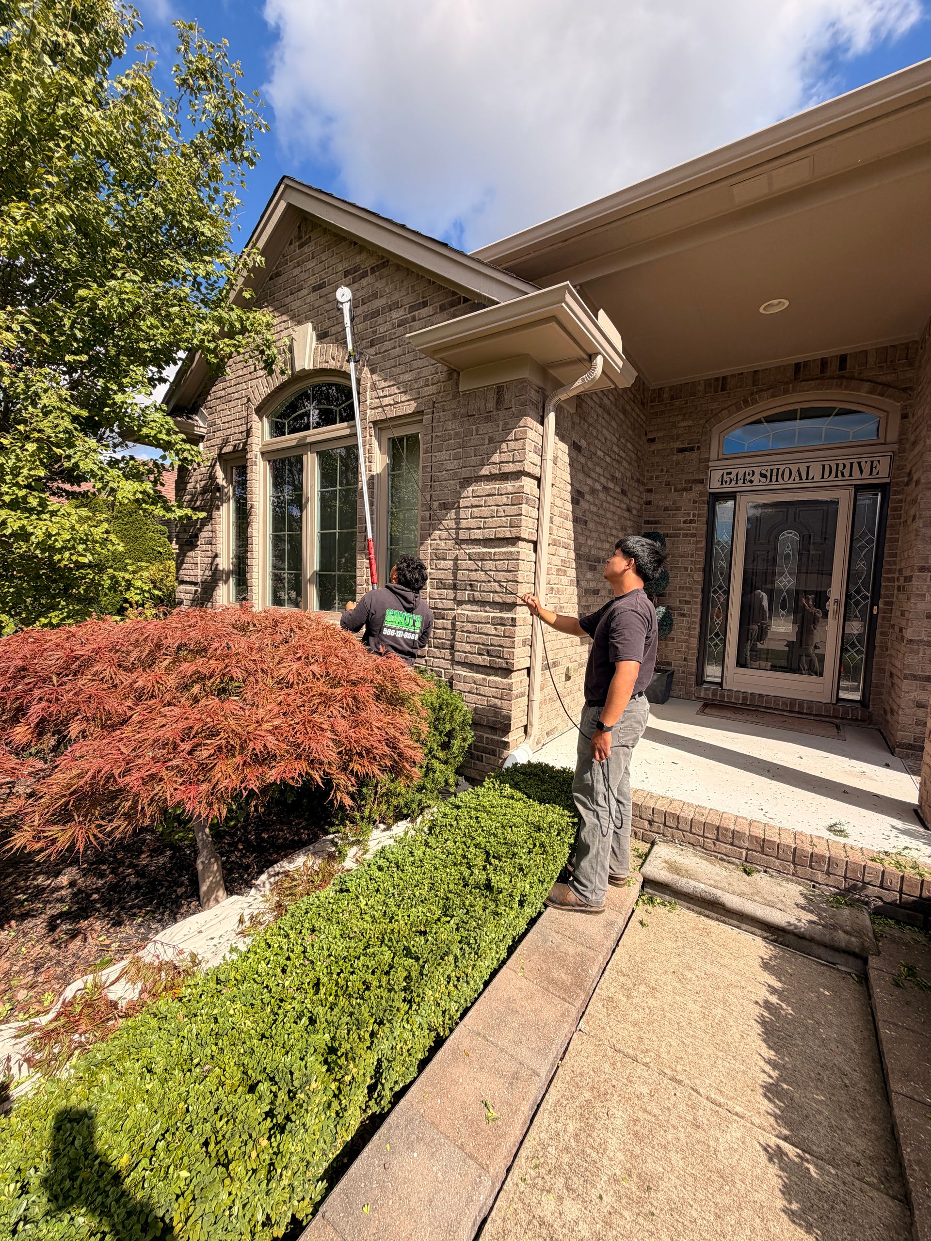 Two people standing by a stone house entrance with arched windows, shrubs, and a sunny walkway