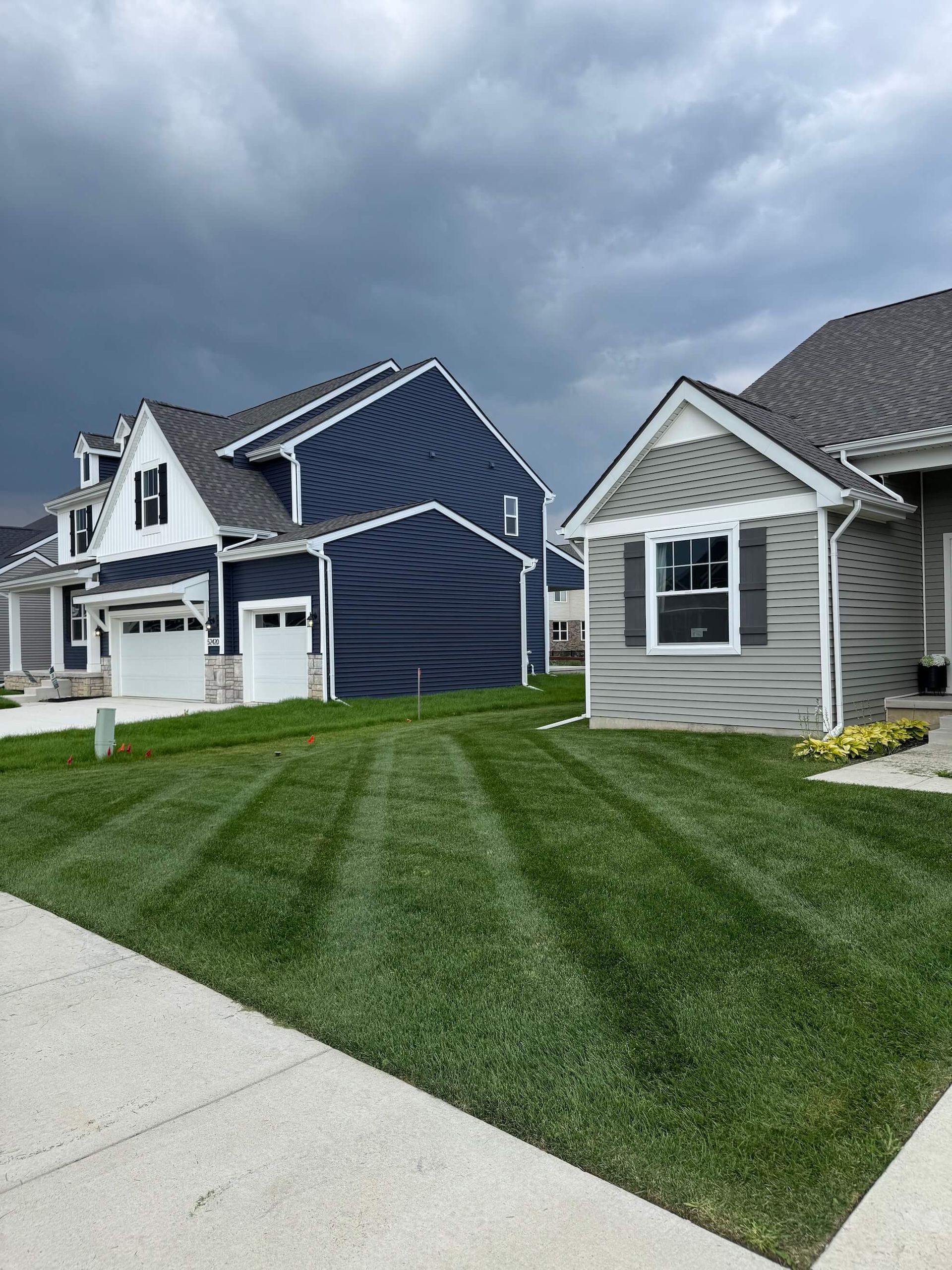 Lawn with striped pattern in front of two houses, one blue and one gray, under a cloudy sky.
