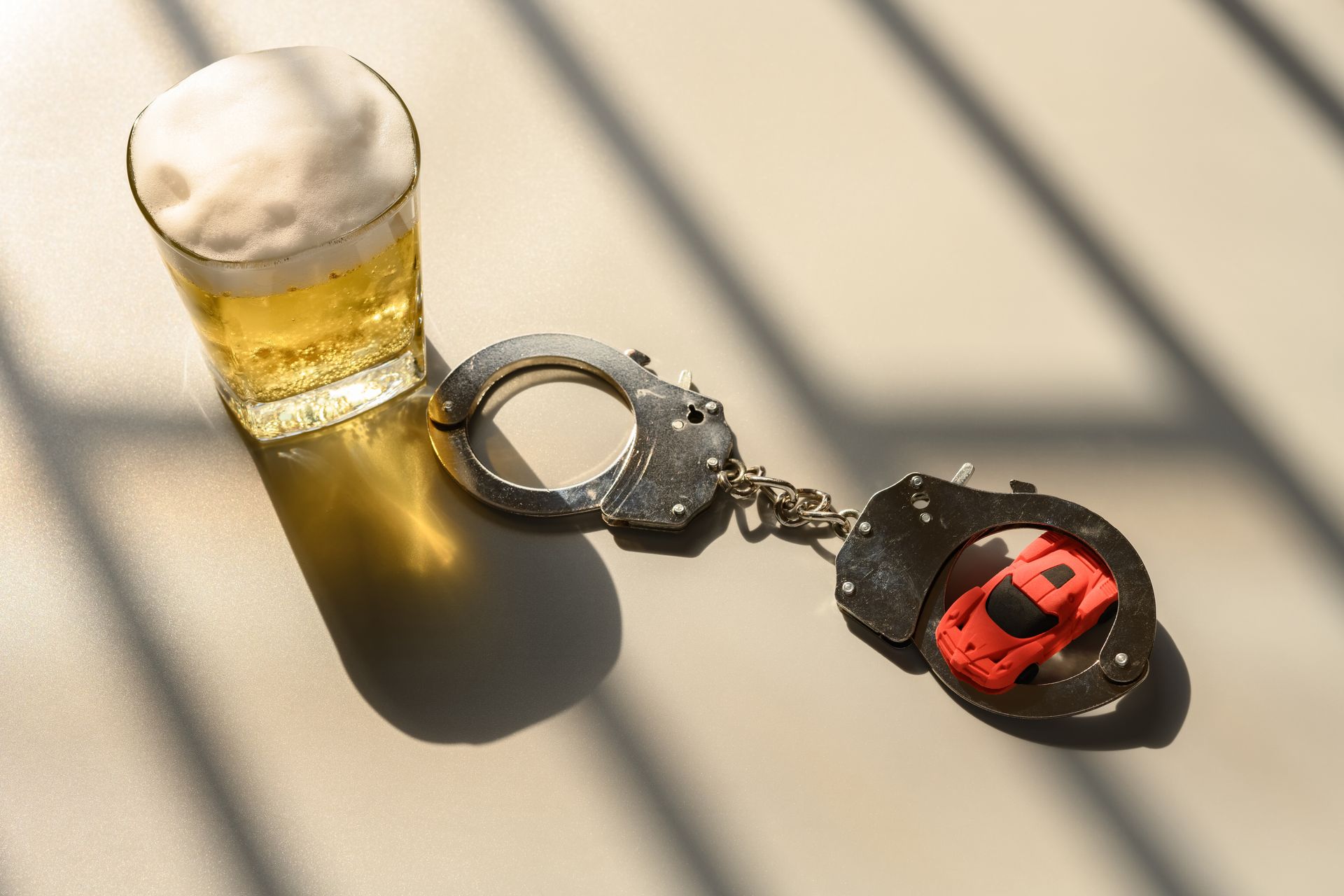 Glass of beer, handcuffs with a toy car, and shadows on a light surface, implying drunk driving.