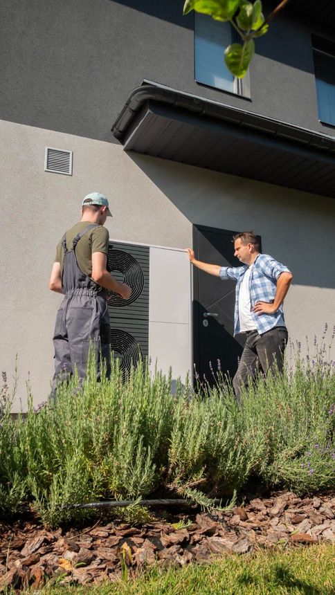 Two workers discussing a house exterior, one pointing at the wall beneath a balcony