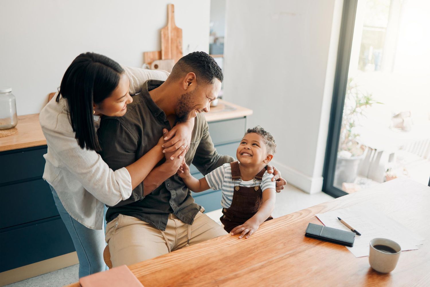 Family sharing a joyful moment at a wooden table in a bright kitchen, with a baby reaching toward an adult.