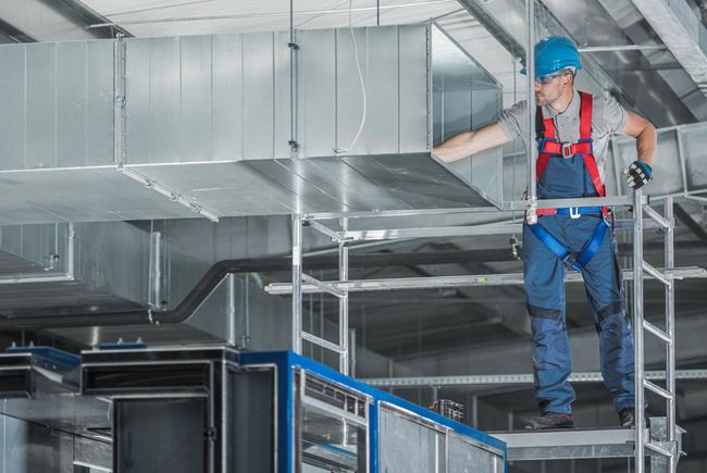 Worker in blue hard hat and harness inspecting ductwork on scaffolding in an industrial ceiling space