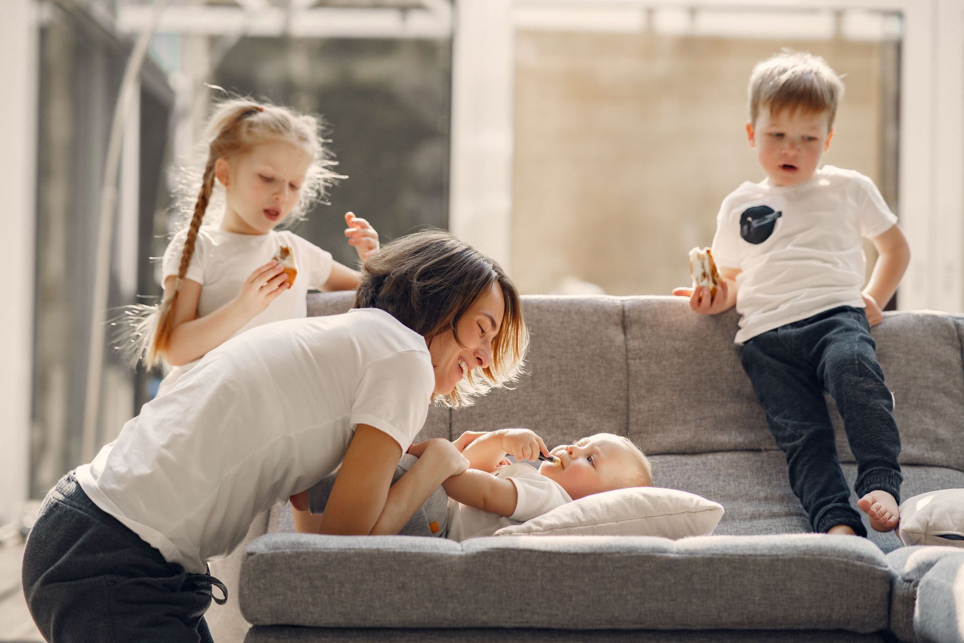 Three children on a gray couch, one leaning over a baby in a bright living room