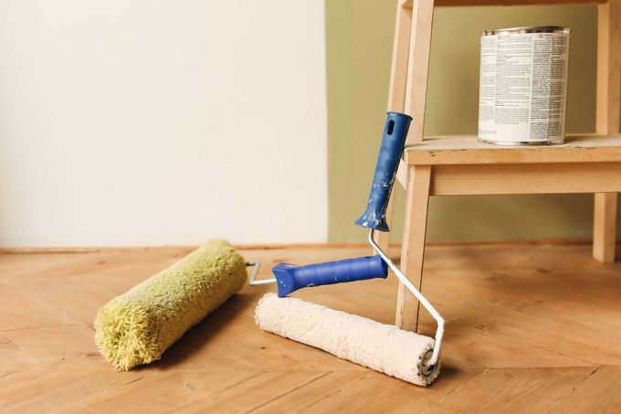 Attrezzi per dipingere su un pavimento di legno: tre rulli e un barattolo di vernice accanto a uno sgabello.