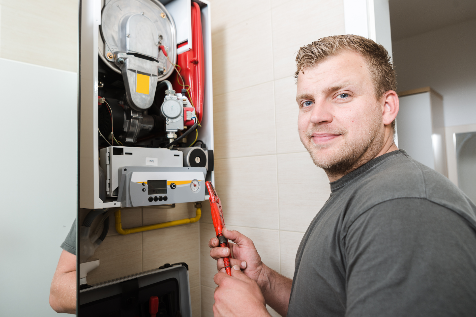 Technician repairing an open wall-mounted boiler with tools in a tiled utility room