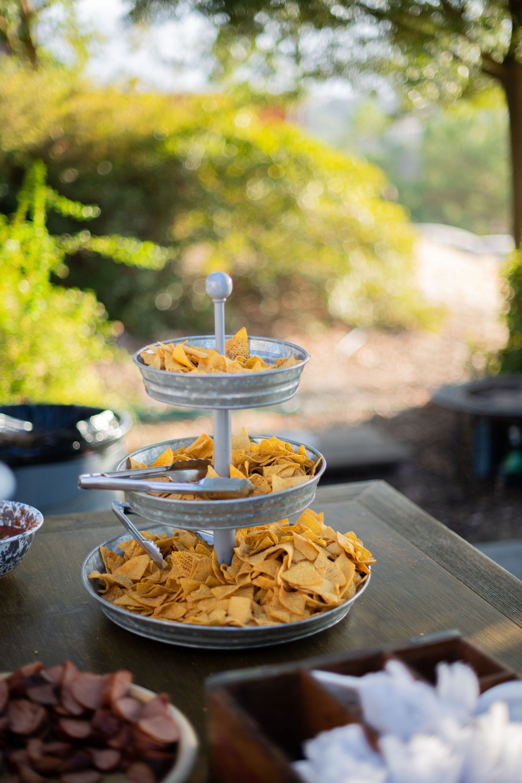 Three-tiered serving dish with tortilla chips outdoors on a table; salsa and other food items nearby.