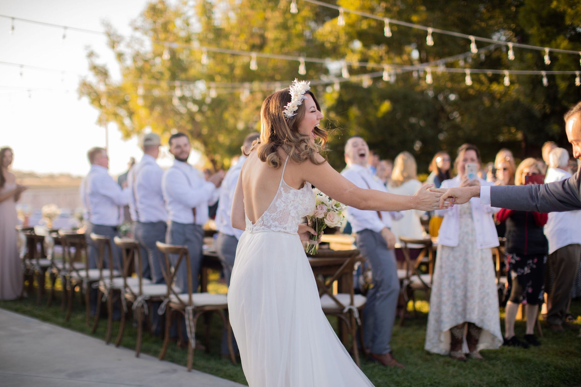 Bride dancing outdoors, holding hands with a person, celebrating. Guests and tables are in the background.