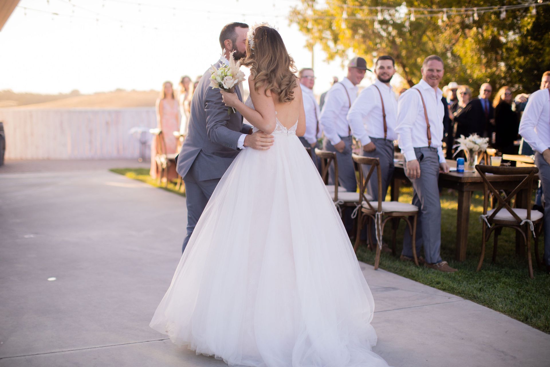 Newlyweds share a kiss during an outdoor wedding ceremony; a wedding party observes in the background.