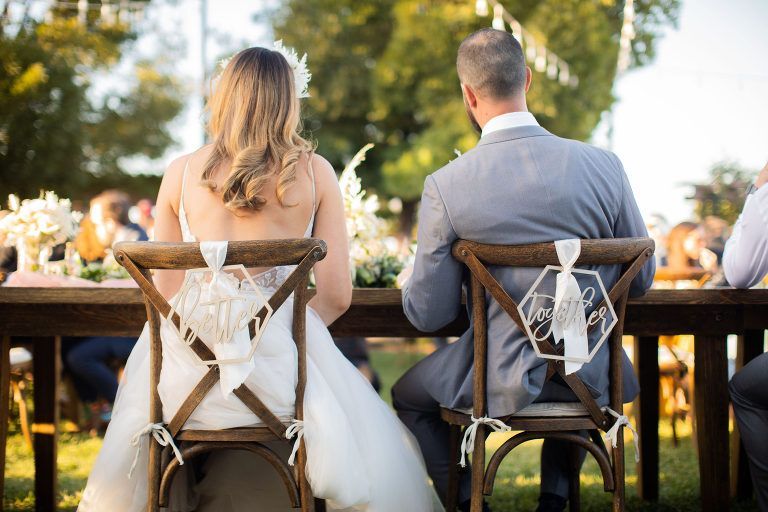 Bride and groom seated at a wooden table outdoors. They are at a wedding reception.