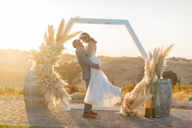 Couple embraces under white arch with pampas grass during golden hour, vineyard in background.