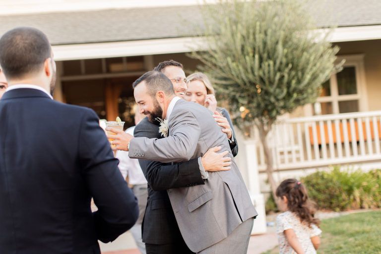 Men in suits embrace, others watch, a wedding celebration outdoors.
