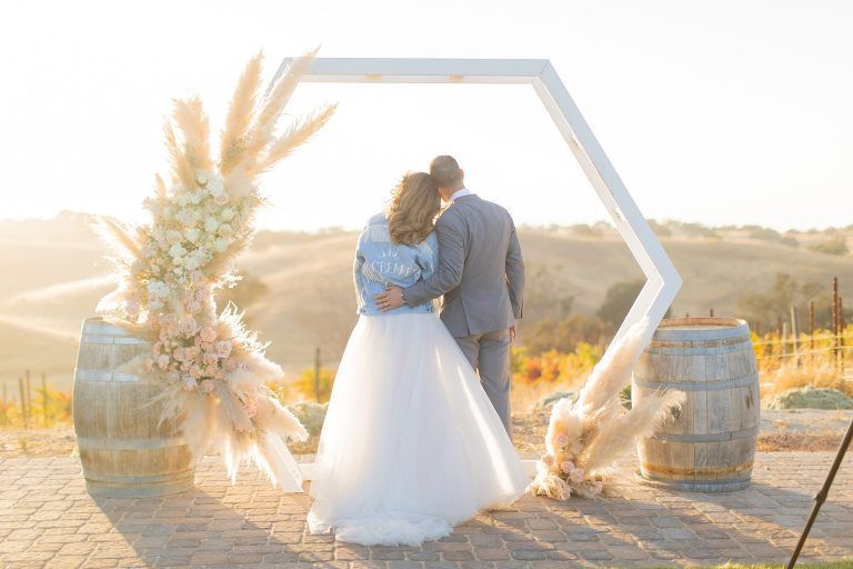 Couple embracing under a white hexagon arch decorated with flowers and pampas grass at an outdoor wedding.