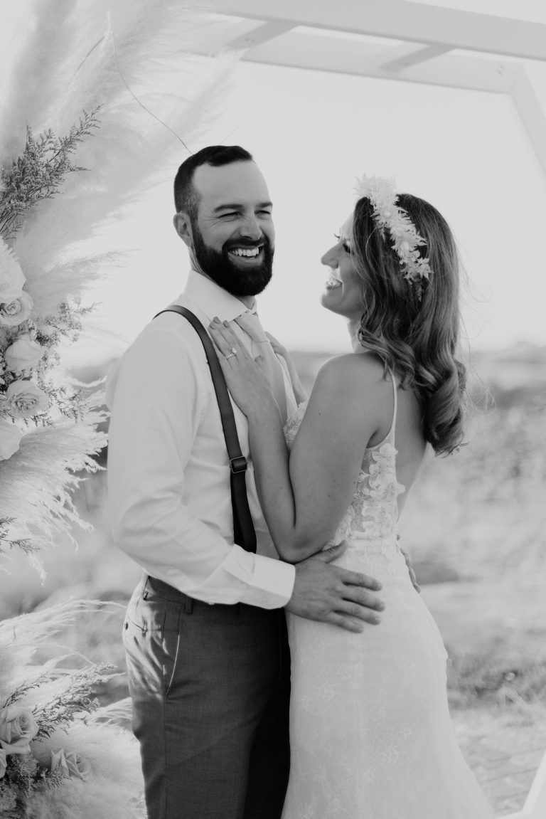 Bride and groom laugh, embracing at an outdoor wedding ceremony. The groom wears suspenders.