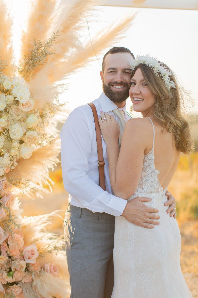 Couple embracing, smiling at camera near floral arch; outdoor wedding.