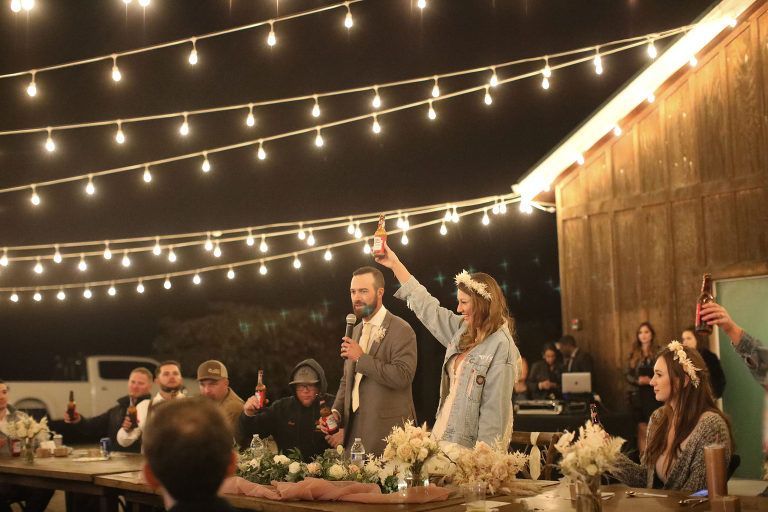 Wedding reception: Couple toasts with beer in front of a barn lit by string lights; guests raise glasses.