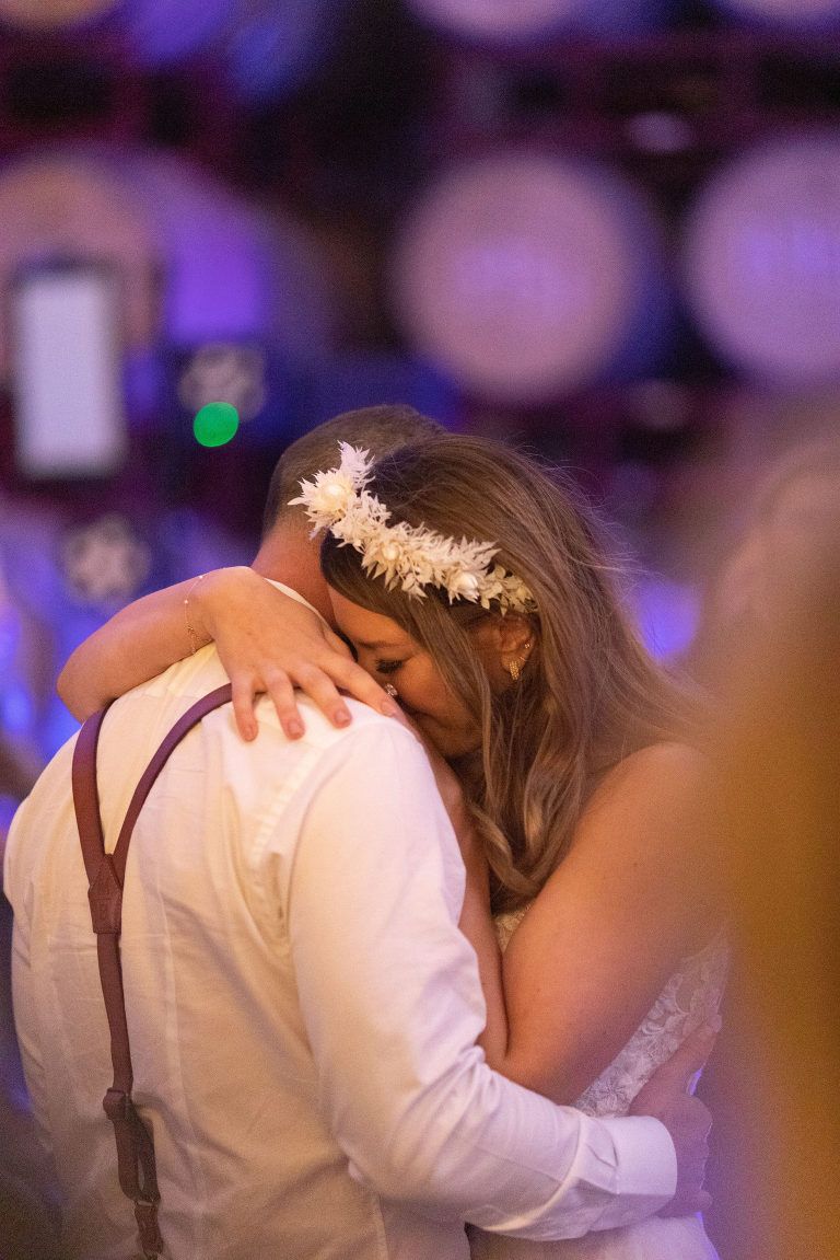Couple dancing, embraced; bride in flower crown, groom in suspenders, soft purple and white background.