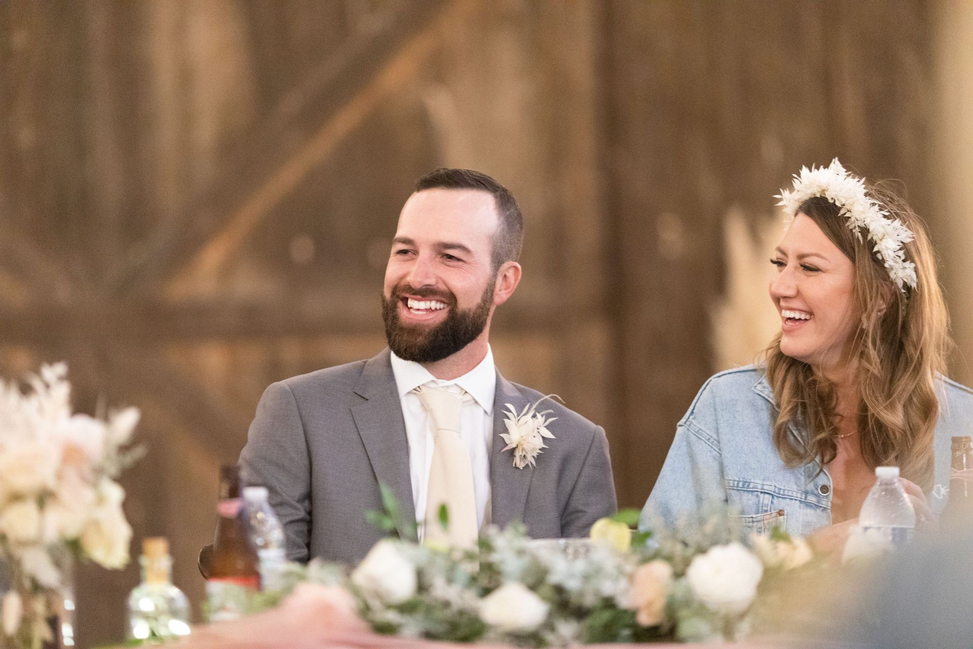 Groom in gray suit laughs, bride in floral crown laughs, seated at a table, floral arrangements, light setting.