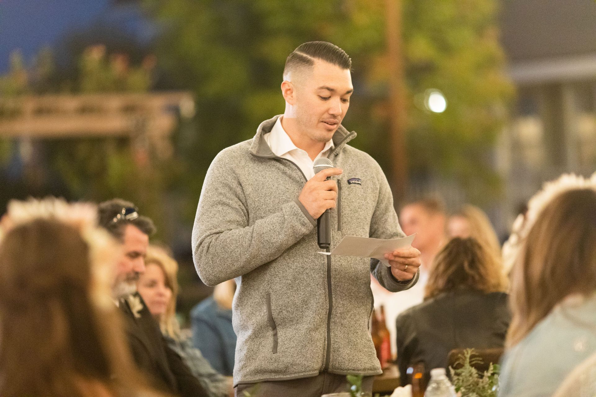 Man speaking at outdoor event, holding microphone and paper. Wearing gray sweater, other people visible.