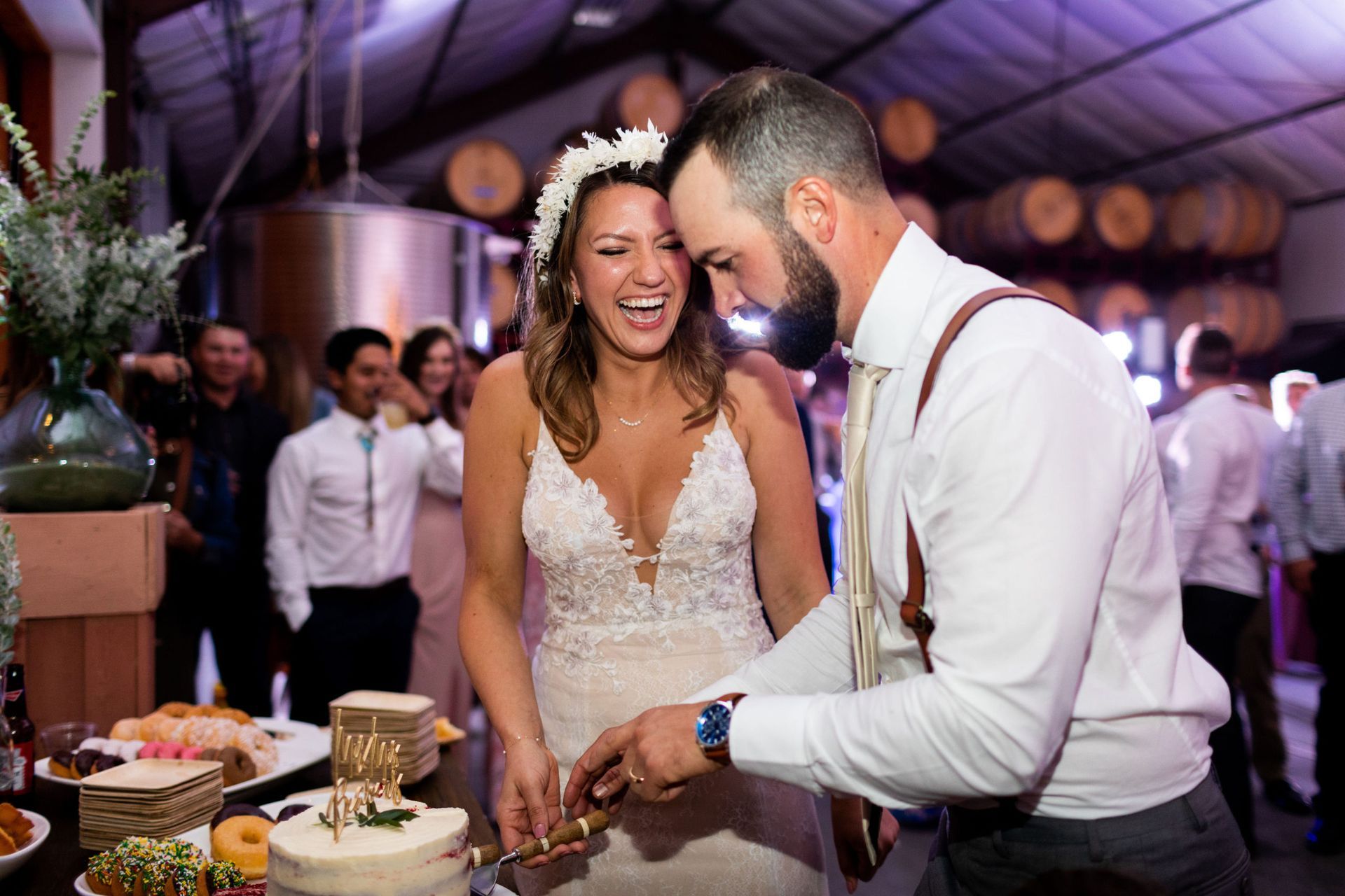 Bride and groom cut wedding cake. Bride laughs, wearing a floral crown and white dress. Groom in suspenders, white shirt.
