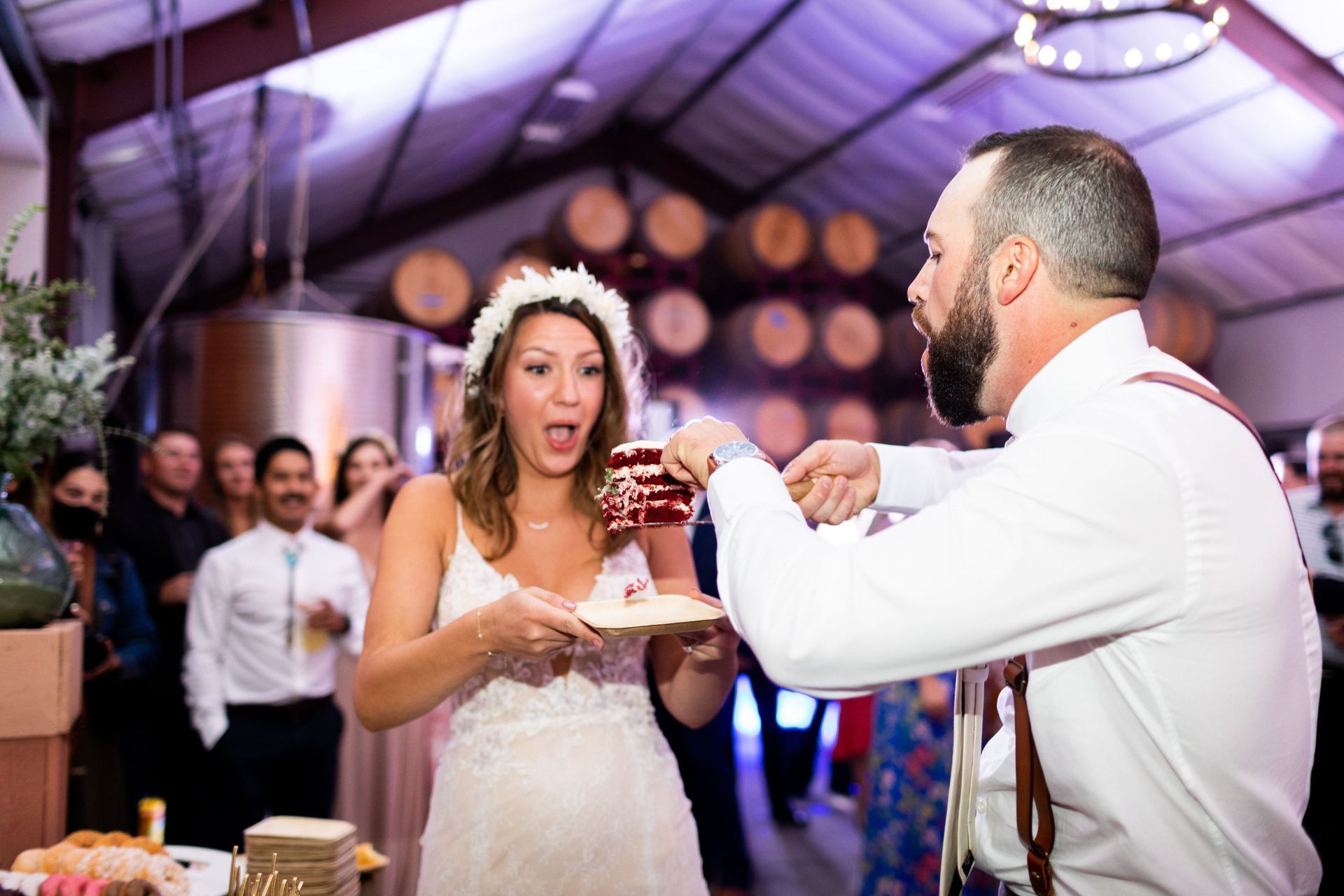 Bride and groom share cake at reception. Bride reacts with surprise. Guests and barrels are in background.