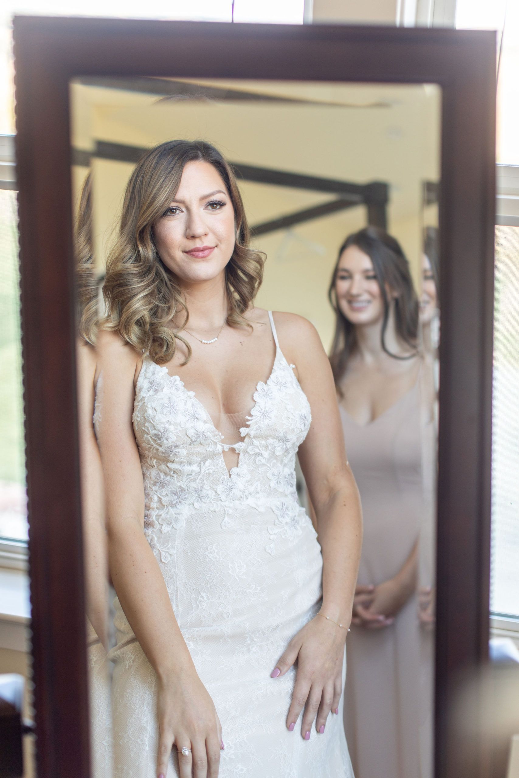 Bride in a white lace dress looks in mirror; bridesmaid smiles behind her.