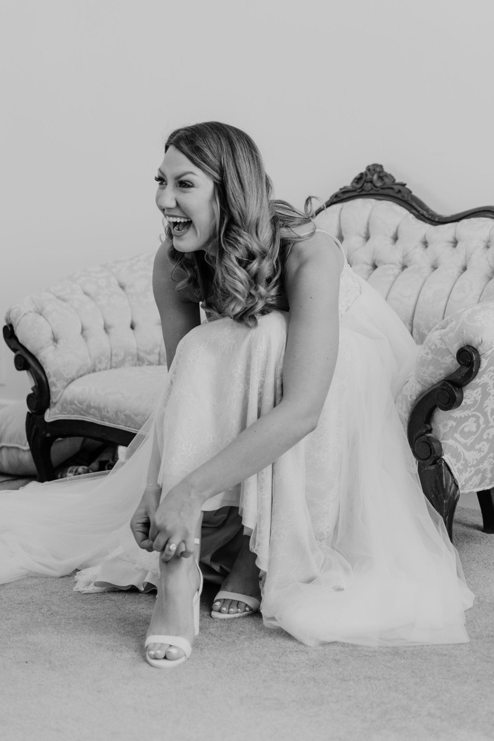 Bride laughing, putting on her wedding shoes, seated on a vintage white sofa.