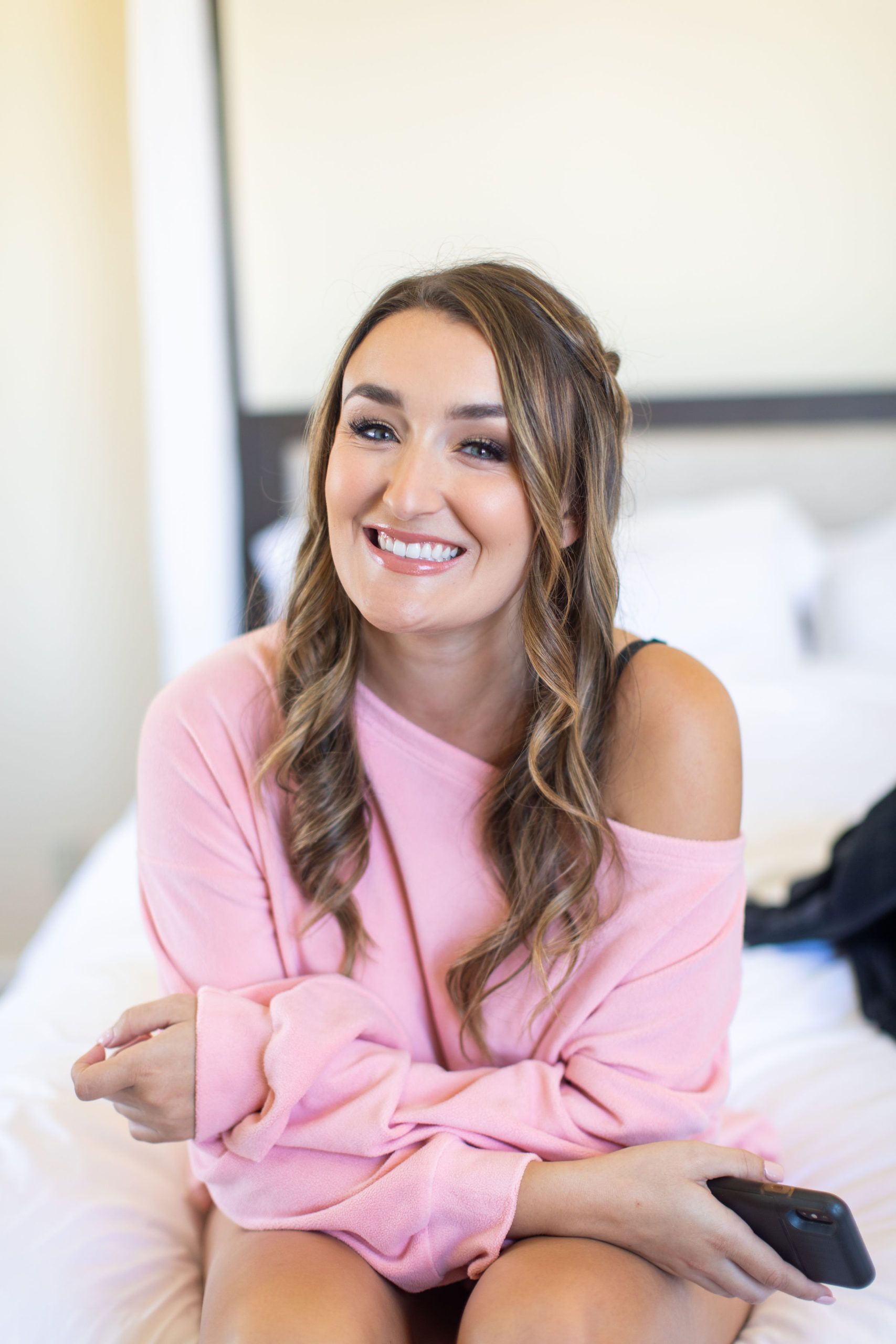 Woman in pink sweater smiling, sitting on a bed.