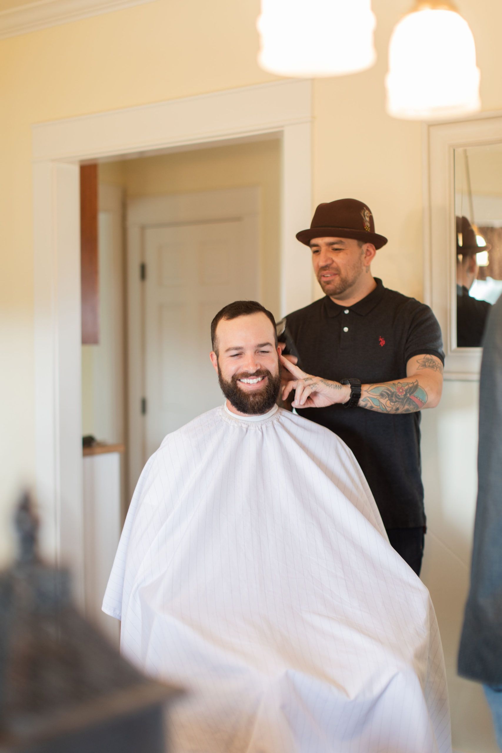 Man getting a haircut in a room with the barber wearing a brown hat and black shirt; white cape; and light colored walls.