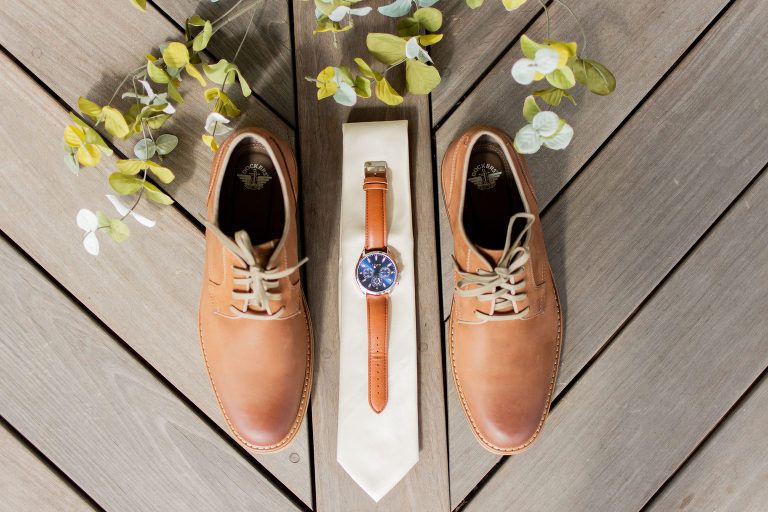 Tan leather shoes, watch, and tie arranged on a wooden surface with greenery.