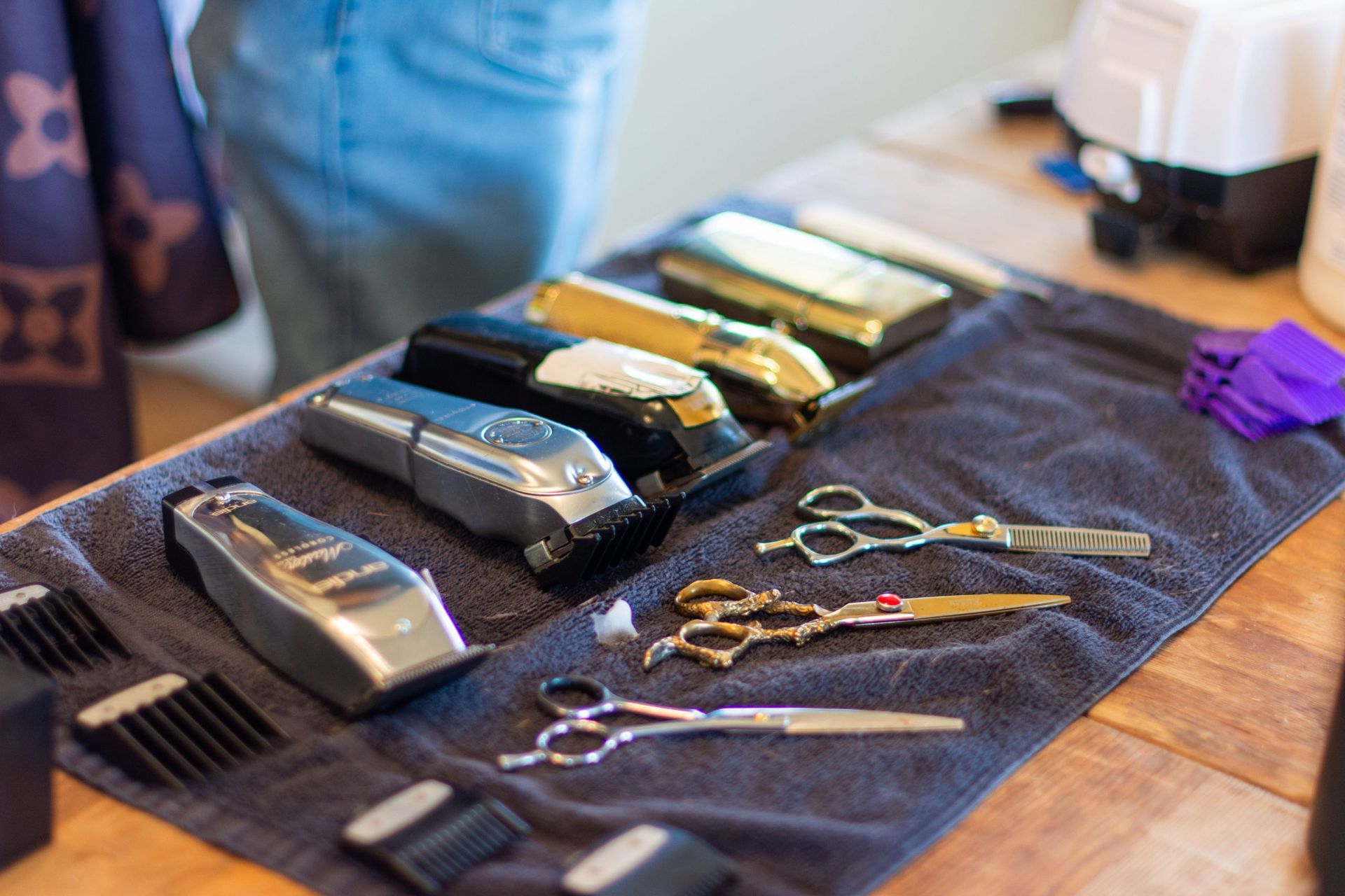 Barber tools, including clippers and scissors, laid out on a dark cloth on a wooden table.
