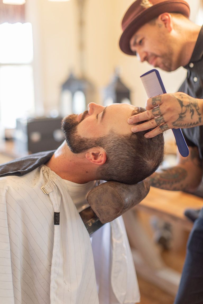 Barber combing customer's hair in a barber shop. Customer has beard, barber wears hat and has tattoos.