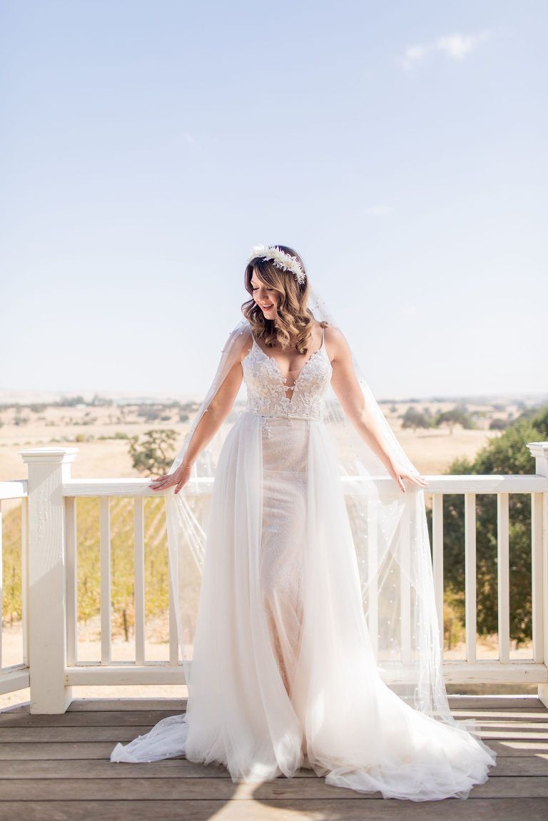 Bride in a white wedding dress with a long veil, standing on a white porch overlooking a landscape.
