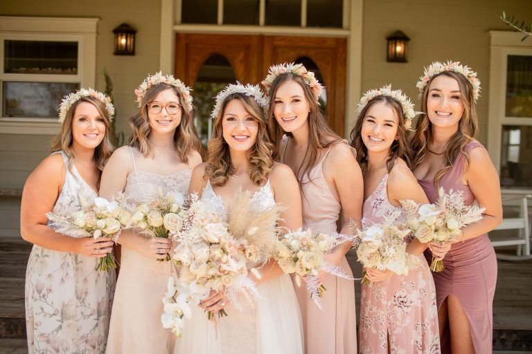 Bridesmaids and bride pose in front of a building. They wear flower crowns and hold bouquets; the dresses are various shades of pink.