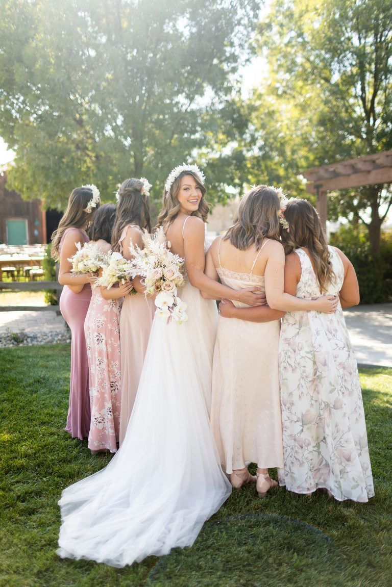 Bride smiles with bridesmaids, posing on grass. Dresses vary in pinks and beige. Sunlight, outdoor setting.