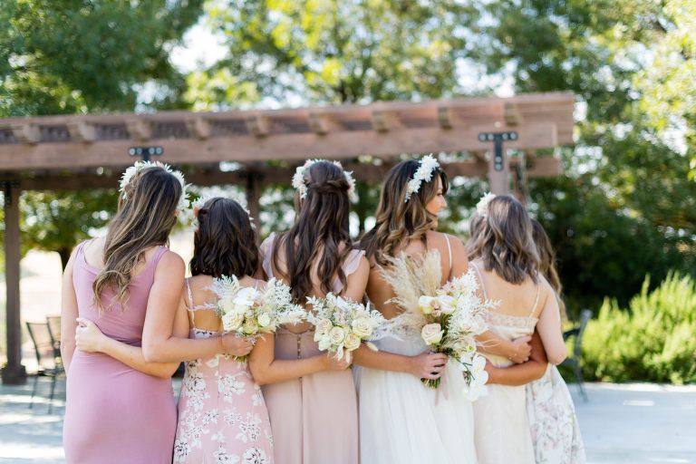 Bridesmaids with bouquets, arms around each other, pose outdoors near a pergola, wearing different pastel dresses.