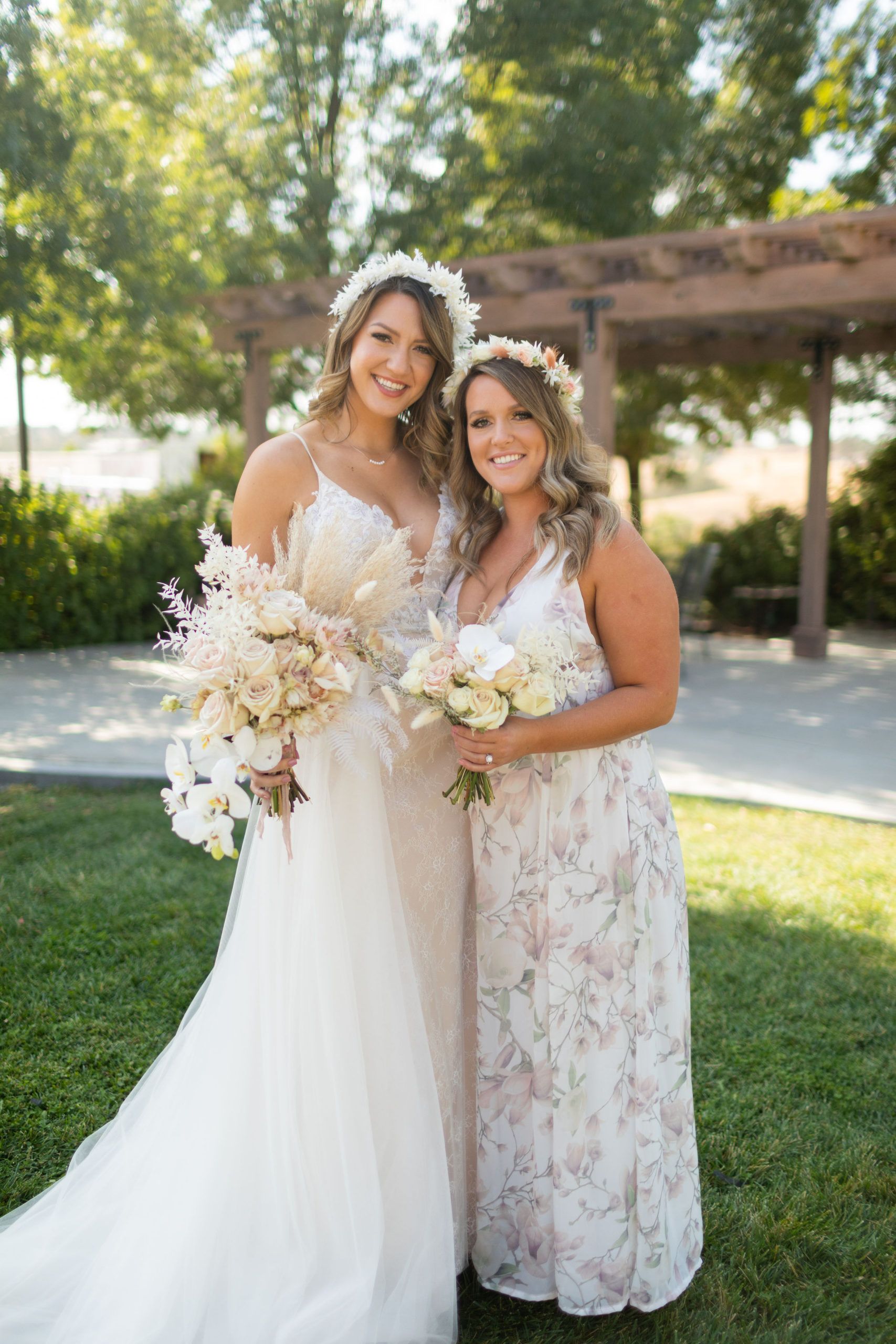 Bride and bridesmaid smiling, holding bouquets, wearing flower crowns, outdoors.