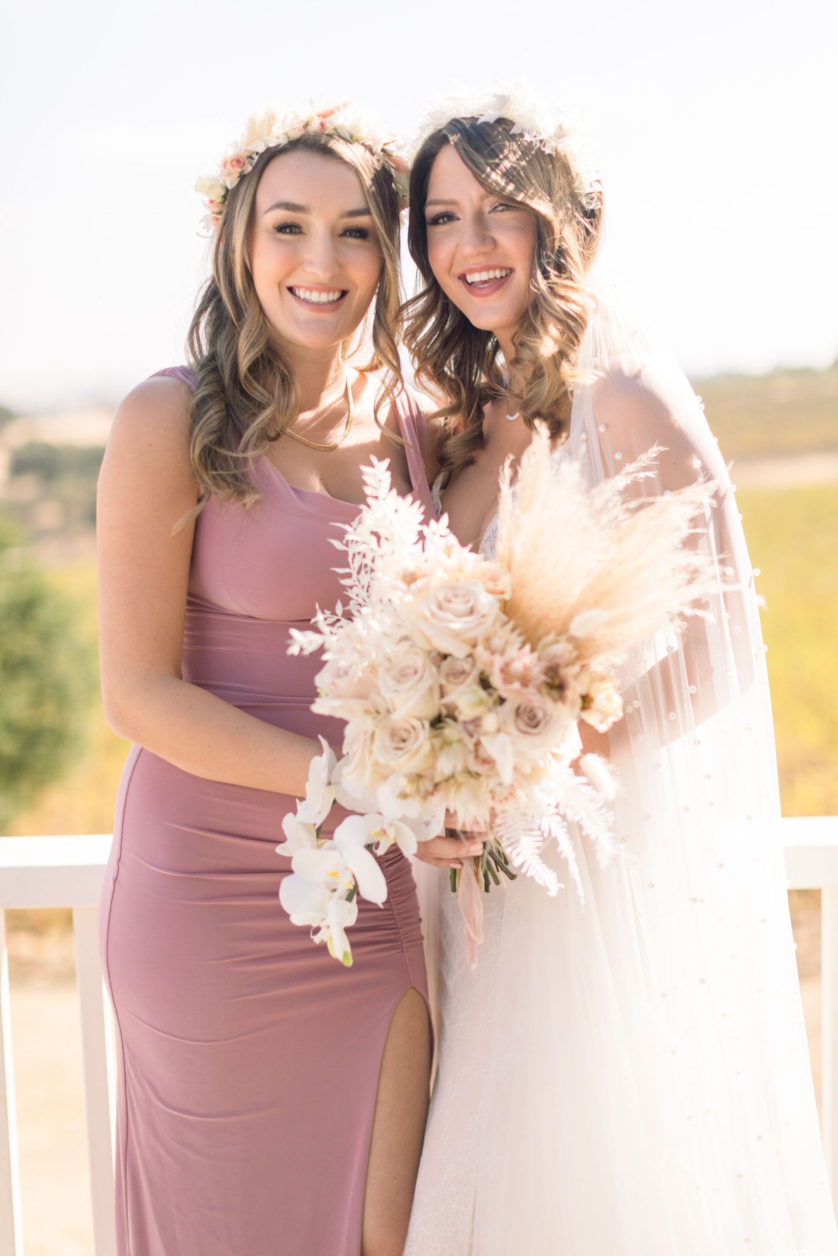 Bride and bridesmaid smiling, holding bouquet, in front of a vineyard; bridesmaid in pink dress.