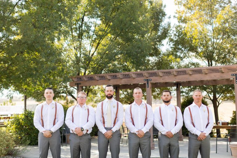 Groomsmen wearing white shirts, gray pants, and suspenders stand outside. Wooden arbor and trees in the background.